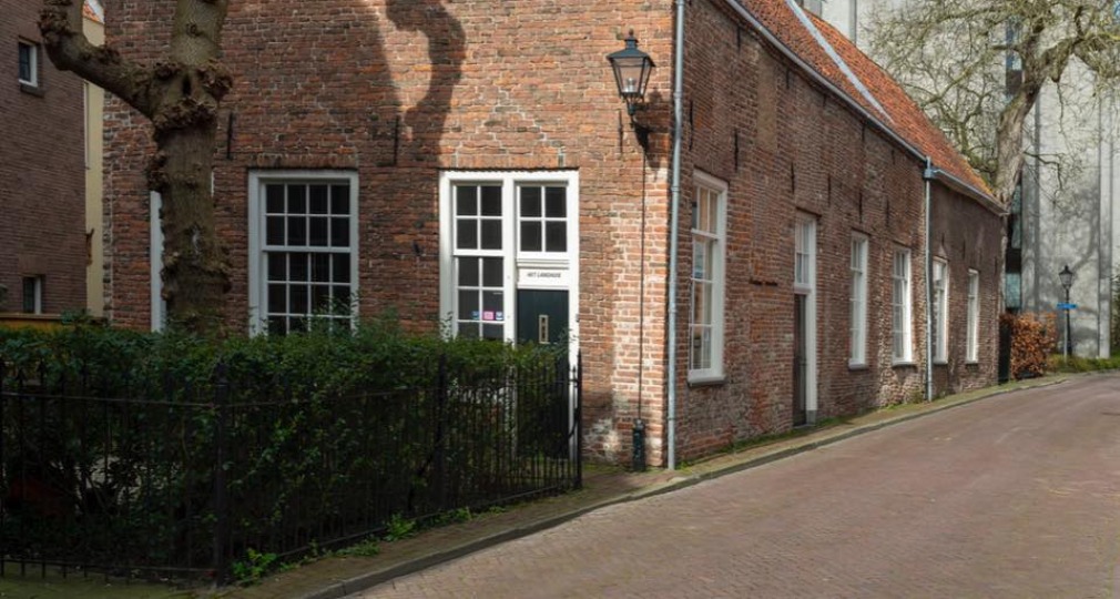 Een historisch bakstenen huis met een rood dak, naast een smalle straat en gesnoeide boom onder blauwe lucht.