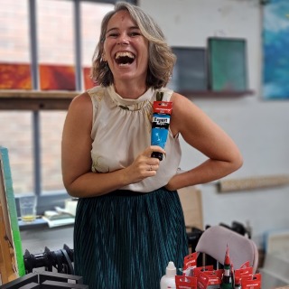 Vrolijke vrouw met schouderlang haar houdt een tube blauwe Amsterdam Expert Acrylverf in een atelier.