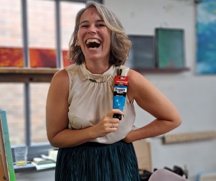 Vrolijke vrouw met schouderlang haar houdt een tube blauwe Amsterdam Expert Acrylverf in een atelier.