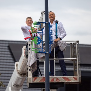 Twee mannen onthullen samen een kleurig straatnaambord terwijl ze op een hoogwerker staan.
