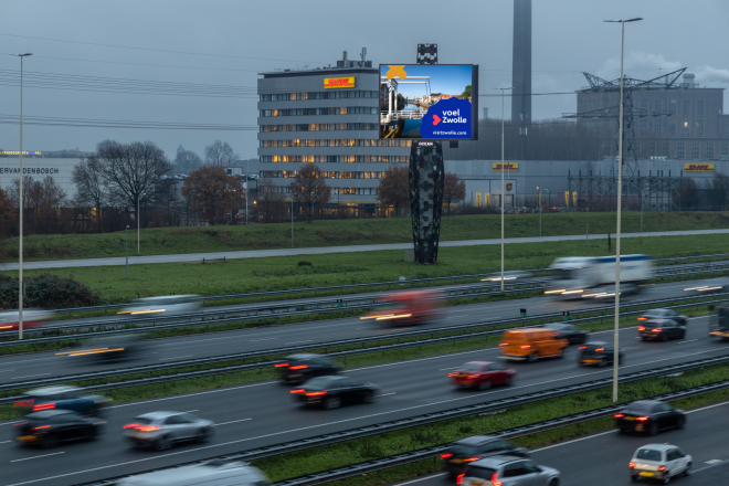 Drukke snelweg bij avond met verlichte reclamebord en DHL-gebouw op de achtergrond in Nederland.