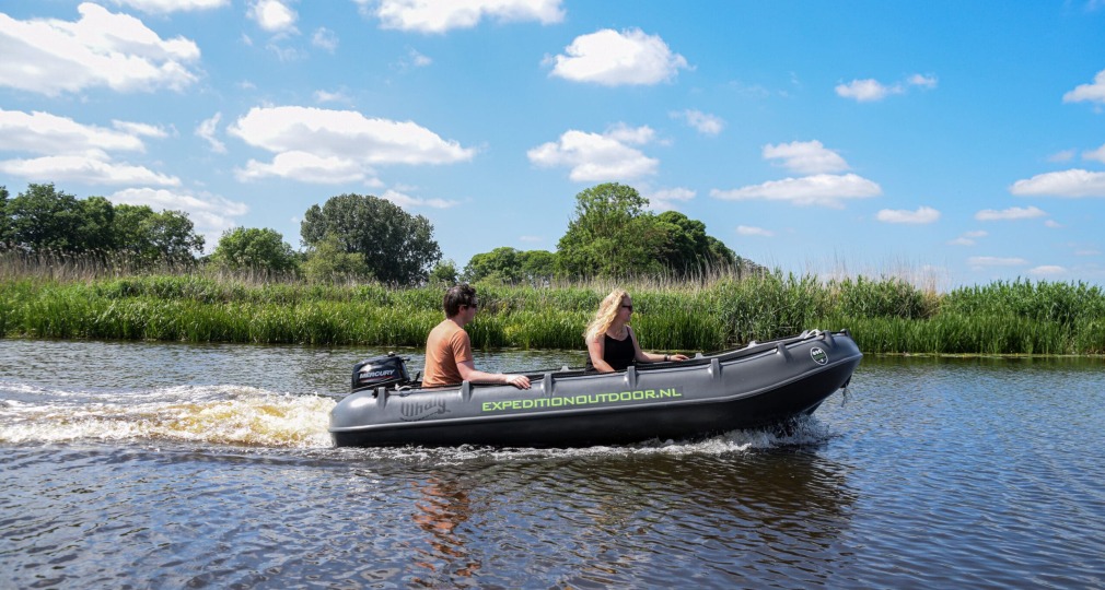 Twee mensen varen in een opblaasbare motorboot op een rustige rivier onder een blauwe lucht met wolken.