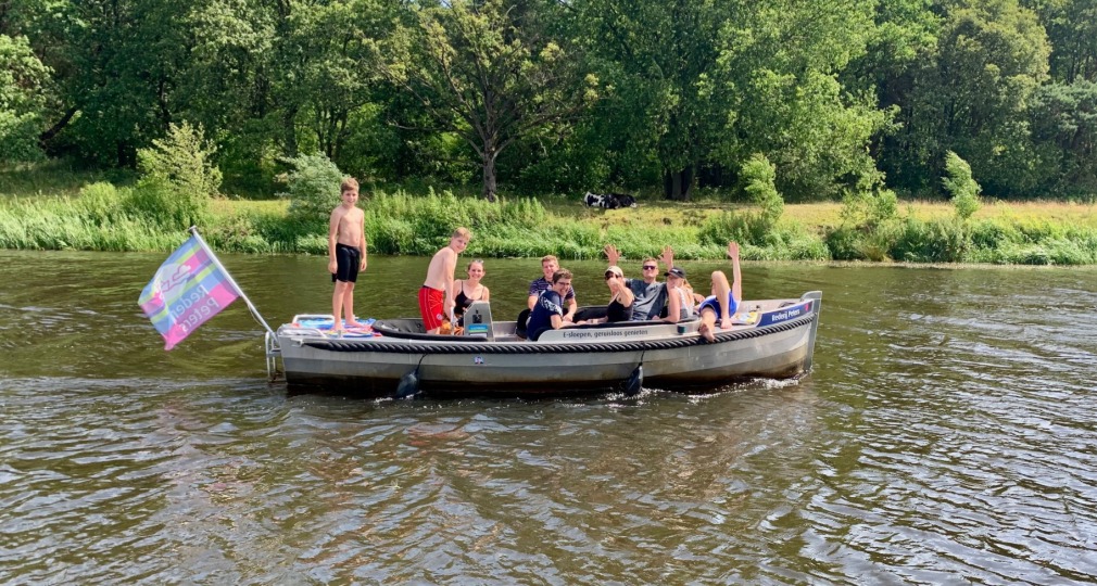 Groep mensen zit in een boot op een rivier, sommigen zwaaien, met kleurrijke vlag en groene bomen op de achtergrond.