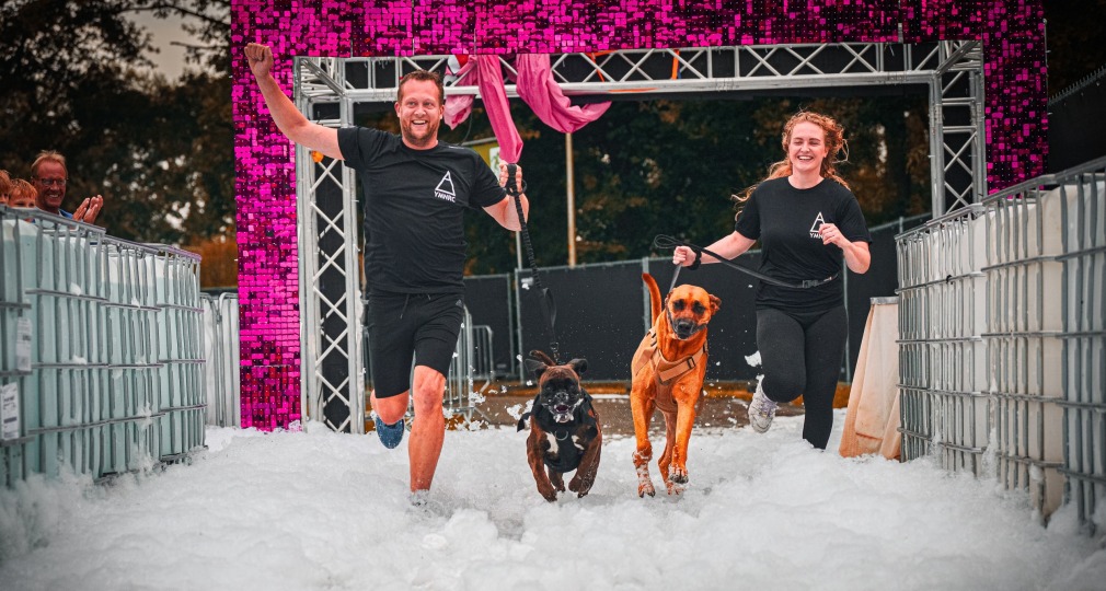 Twee mensen rennen met hun honden door een schuimbad tijdens een vrolijk sportevenement buiten.
