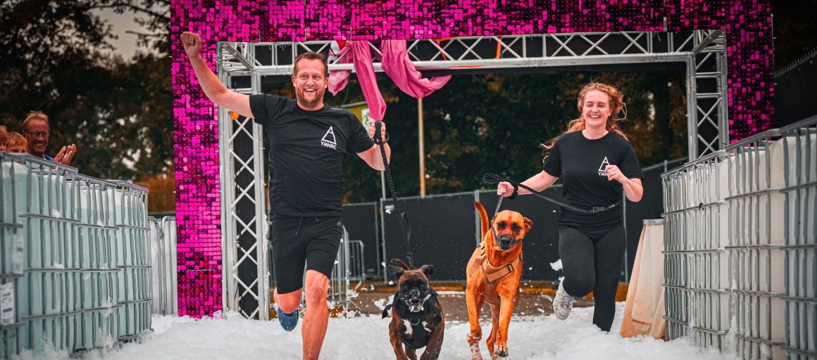 Twee mensen rennen met hun honden door een schuimbad tijdens een vrolijk sportevenement buiten.