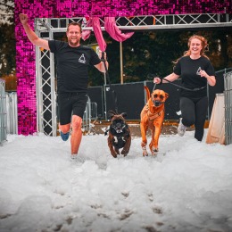 Twee mensen rennen met hun honden door een schuimbad tijdens een vrolijk sportevenement buiten.