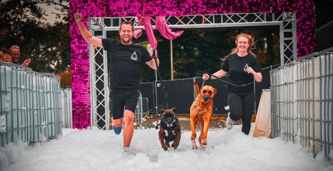 Twee mensen rennen met hun honden door een schuimbad tijdens een vrolijk sportevenement buiten.