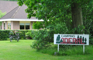 Camping IDEAL-bord bij een huis met tuin, bloemenkar, en groene bomen in een landelijke omgeving.