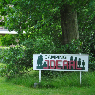 Camping IDEAL-bord bij een huis met tuin, bloemenkar, en groene bomen in een landelijke omgeving.