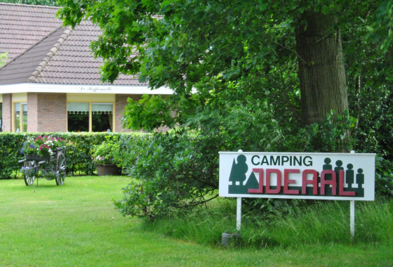Camping IDEAL-bord bij een huis met tuin, bloemenkar, en groene bomen in een landelijke omgeving.