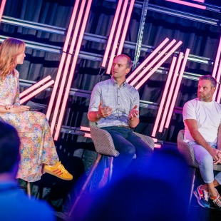 Panelgesprek met drie personen op een podium met neonverlichting en een publiek op de voorgrond.