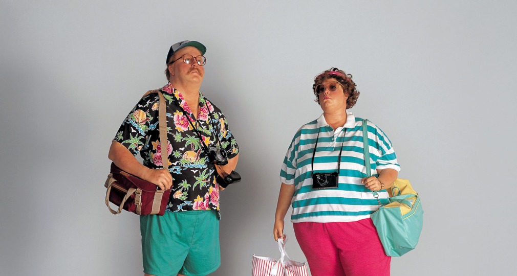 Twee toeristen poseren in kleurrijke, casual kleding met tassen, camera's, en sandalen tegen een grijze muur.