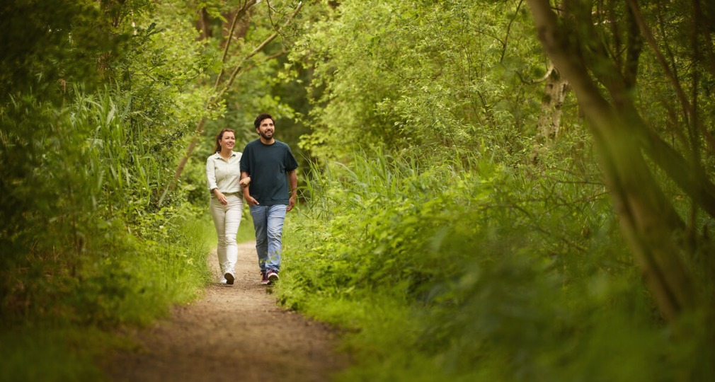 Twee mensen wandelen samen over een bospad, omringd door groene bomen en planten op een zonnige dag.