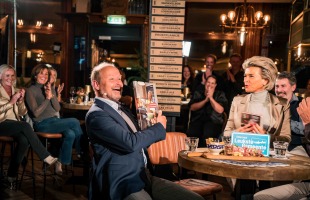 Een man viert een prijs in een gezellige kroegsetting, omringd door applaudisserende mensen aan tafels.