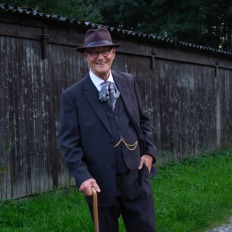 Älterer Mann im dunklen Anzug mit Hut und Stock, lächelt stehend neben einem alten Holzschuppen.
