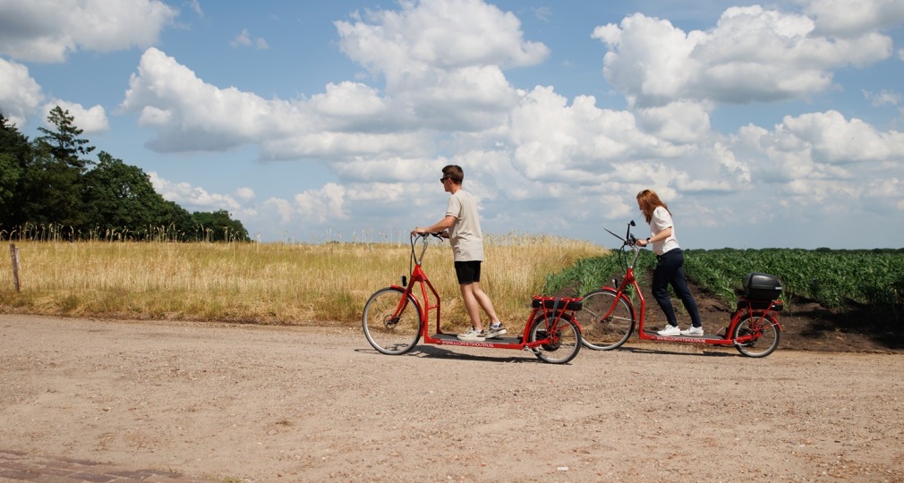 Twee mensen rijden op rode elektrische steppen langs een landweg met velden en blauwe lucht met wolken.