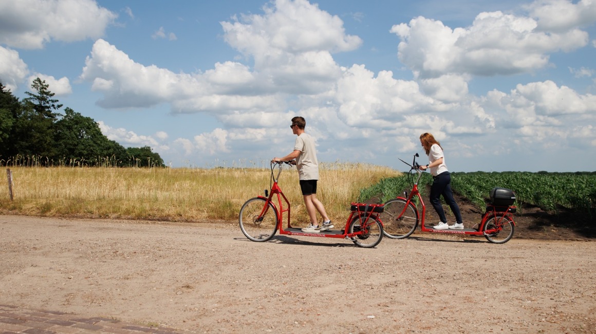 Twee mensen rijden op rode elektrische steppen langs een landweg met velden en blauwe lucht met wolken.
