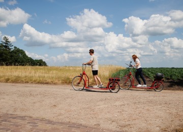 Twee mensen rijden op rode elektrische steppen langs een landweg met velden en blauwe lucht met wolken.