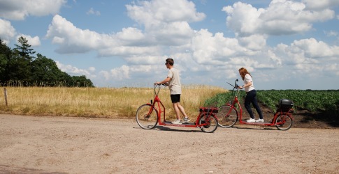 Twee mensen rijden op rode elektrische steppen langs een landweg met velden en blauwe lucht met wolken.