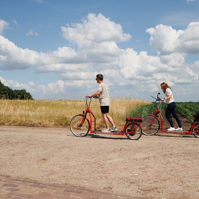 Twee mensen rijden op rode elektrische steppen langs een landweg met velden en blauwe lucht met wolken.