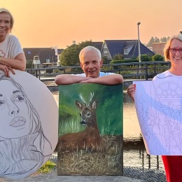 Drie mensen poseren buiten met kunstwerken: een portret, een hertenschilderij en een lijntekening.