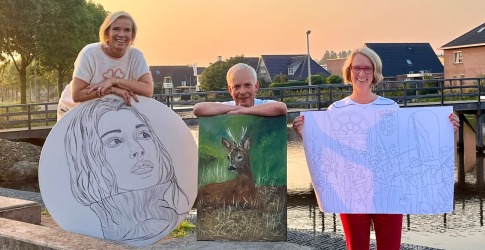 Drie mensen poseren buiten met kunstwerken: een portret, een hertenschilderij en een lijntekening.