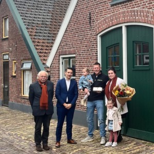 Een familie en twee mannen poseren voor een traditioneel huis met groene deuren en bakstenen muren.