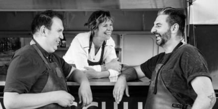 Drie lachende mensen, twee mannen met schorten en een vrouw in wit overhemd, praten bij een kraam.