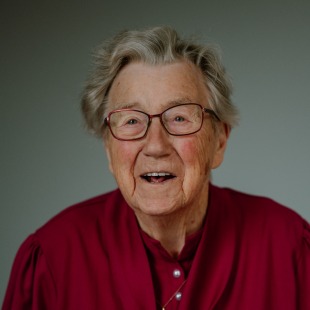 Oudere vrouw met grijs haar en bril draagt een bordeauxrode blouse en glimlacht vriendelijk naar de camera.