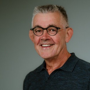 Portret van een oudere man met grijs haar en bril, lachend naar de camera in een donker poloshirt.