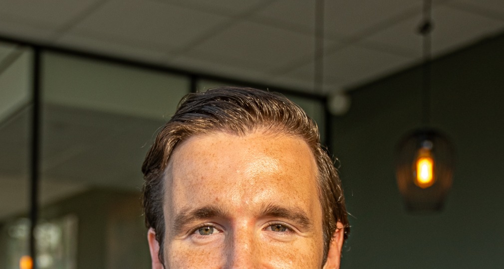 Man met kort bruin haar in een groen T-shirt, glimlachend zittend in een modern kantoorinterieur.