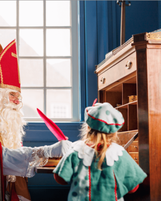 Sinterklaas met rode mijter en staf praat met een meisje verkleed als Piet in een traditioneel kantoor.