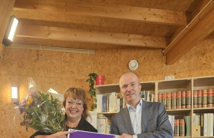 Twee mensen poseren binnen; de vrouw houdt bloemen en een groot certificaat met de naam Judith Elders.