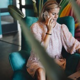 Vrouw in roze jurk werkt op laptop en praat aan de telefoon, zittend op een groene stoel in een modern interieur.