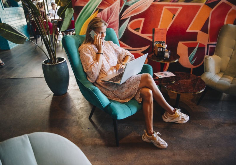 Vrouw zit in een moderne lounge, werkt op een laptop en telefoont in een kleurrijk café, graffiti op muur.