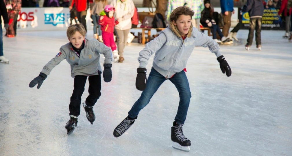 Twee kinderen schaatsen samen op een drukke ijsbaan, lachend en gekleed in winterjassen en handschoenen.