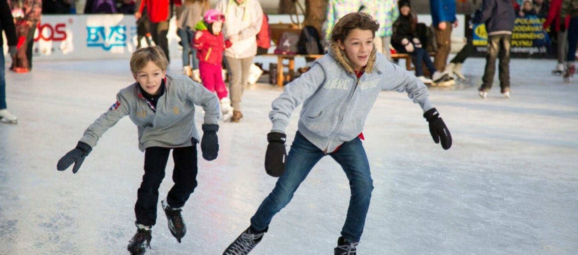 Twee kinderen schaatsen samen op een drukke ijsbaan, lachend en gekleed in winterjassen en handschoenen.