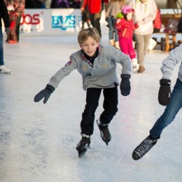 Twee kinderen schaatsen samen op een drukke ijsbaan, lachend en gekleed in winterjassen en handschoenen.