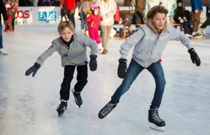 Twee kinderen schaatsen samen op een drukke ijsbaan, lachend en gekleed in winterjassen en handschoenen.