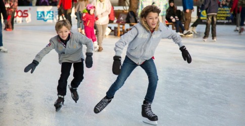 Twee kinderen schaatsen samen op een drukke ijsbaan, lachend en gekleed in winterjassen en handschoenen.