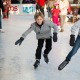 Twee kinderen schaatsen samen op een drukke ijsbaan, lachend en gekleed in winterjassen en handschoenen.