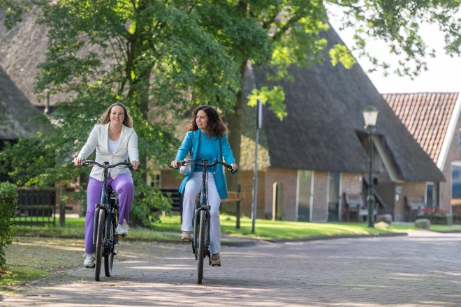 Twee vrouwen fietsen samen op een rustige, groene straat langs traditionele Nederlandse huizen.