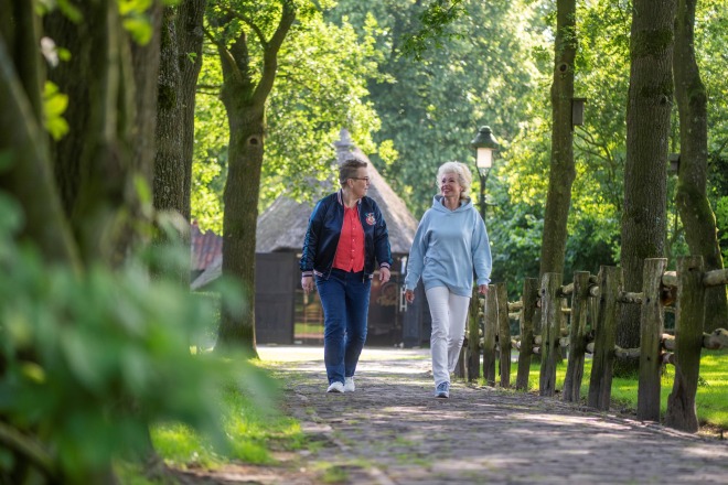 Twee oudere vrouwen wandelen samen op een zonnig, groen bospad met houten hek en bomen.