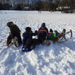 Vier kinderen zitten met sleeën in de sneeuw, spelend op een besneeuwd veld met bomen en huizen.