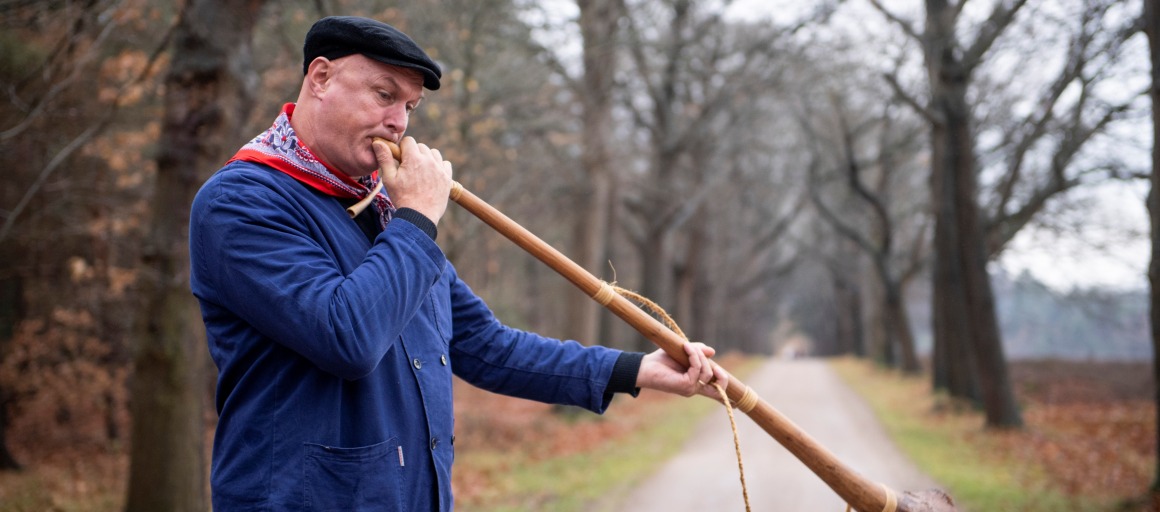 Man in blauwe jas en zwarte pet blaast op een lange houten hoorn op een landweg omringd door bomen.