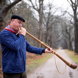 Man in blauwe jas en zwarte pet blaast op een lange houten hoorn op een landweg omringd door bomen.