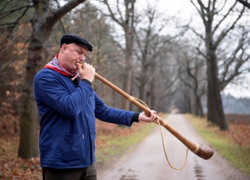 Man in blauwe jas en zwarte pet blaast op een lange houten hoorn op een landweg omringd door bomen.