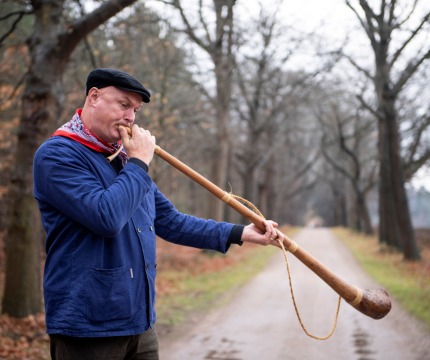Man in blauwe jas en zwarte pet blaast op een lange houten hoorn op een landweg omringd door bomen.