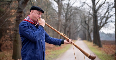 Man in blauwe jas en zwarte pet blaast op een lange houten hoorn op een landweg omringd door bomen.