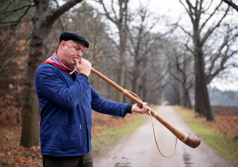 Man in blauwe jas en zwarte pet blaast op een lange houten hoorn op een landweg omringd door bomen.